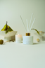 A white reed diffuser bottle with a gold collar and white reeds stands on a white surface beside a matching white candle. The scene is styled with a green pear in a white bowl, a glass bowl with dried botanicals, and a small decorative ball.
