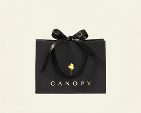 A black CANOPY shopping bag with a black ribbon bow and gold lettering, centered on a light cream background.