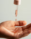 A close-up of a hand holding a small pool of translucent pink liquid under a glass dropper pipette with a pale pink cap, against a soft light gray background.