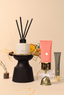 A curated beauty and home gift set featuring a black reed diffuser with white wax, a pink tube of face cream, a gray tube of eye cream, and a small gold bell, styled on a beige background with dried grass and a small vase.
