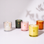 Five lit scented candles in different glass containers arranged on a white surface against a white wall with soft leaf shadows. The candles include clear, dark green, pink, yellow, and amber glass jars, each with a small gold leaf logo and the brand name CANOPY visible on the front.