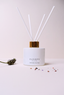 A white reed diffuser bottle with a gold collar and four white reeds, labeled 'CANOPY HOME' and 'LAVENDER & WILD MOSS', on a light beige background with a small lavender sprig and scattered petals nearby.
