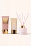 Three Canopy home fragrance products arranged on a warm off-white background: a pink body wash tube with a gold cap, a beige lotion tube with a gold cap, and a pale pink reed diffuser bottle with gold collar and pink reeds.