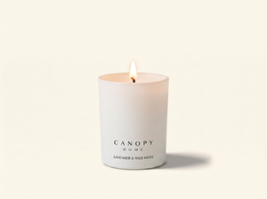 A lit white scented candle in a matte white glass jar with the brand name CANOPY HOME and scent name LAVENDER & WILD MOSS printed on the front, set against a warm off-white background.