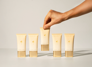 Five CANOFY skincare tubes in a warm cream color with gold caps are lined up on a white surface, with a hand reaching to pick up the center tube against a clean white background.
