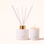 A reed diffuser bottle with white reeds and a gold collar stands beside a matching white candle in a glass holder with a small flame. Both items are labeled CANOPY HOME and FRAGRANCE OF & MELT, on a warm off-white background.