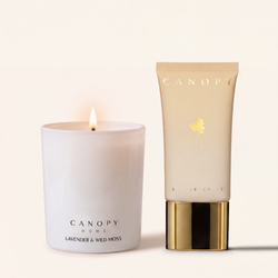 A lit white candle and a beige squeeze tube of CANOPY Home Skincare Creme on a warm cream background. The candle is labeled 'LAVENDER & WILD MOSS' and the tube is labeled 'SKIN CARE CREME' with a gold leaf logo.