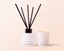 A reed diffuser bottle with black reeds and a matching white candle jar, both labeled CANOPY PARIS Orange, Bergamot & Ember, set against a warm beige background.